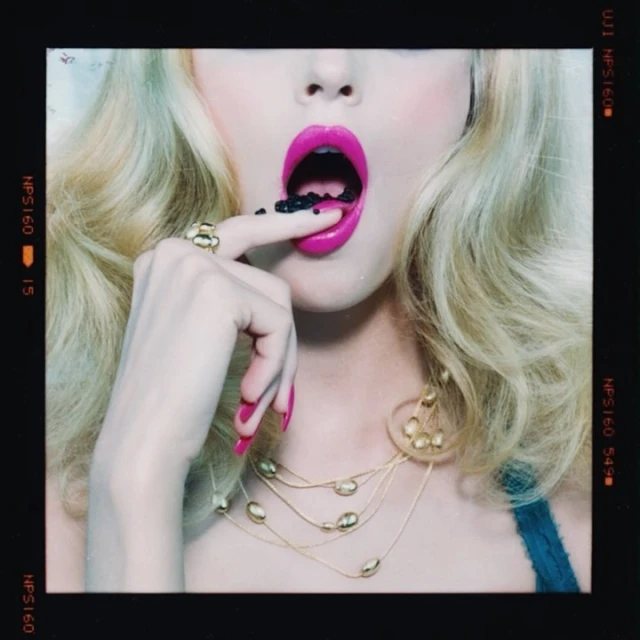 Close-up of a woman with blonde hair, bright pink lipstick, holding small black objects on her finger and near her open mouth.
