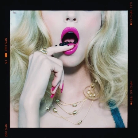 Close-up of a woman with blonde hair, bright pink lipstick, holding small black objects on her finger and near her open mouth.