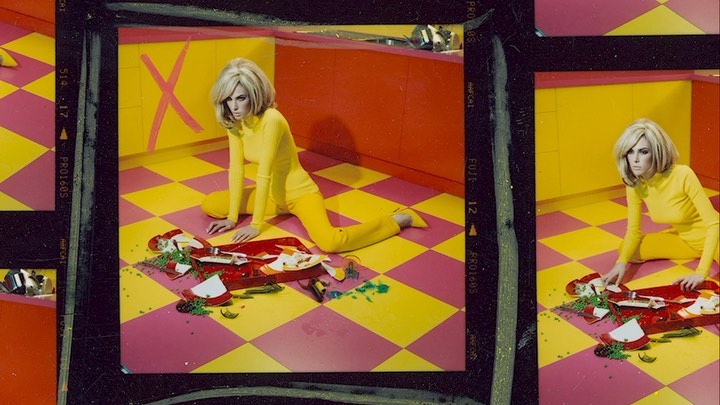 Woman in yellow outfit poses on checkered floor with broken plates in front of orange and yellow kitchen cabinets with X mark.