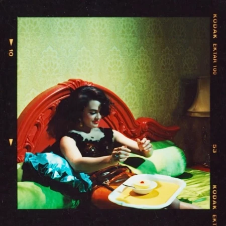 A woman in a black top sits on a green bed with a red headboard holding a small dessert tray.