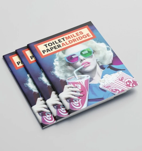 Close-up of three fashion magazines stacked, featuring a woman with retro 3D glasses drinking Coca-Cola and eating popcorn.