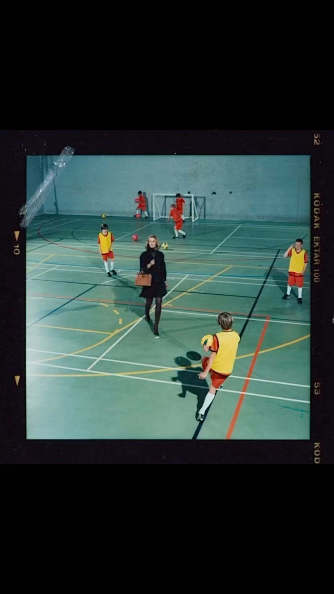 Fashionably dressed adult female stands on a sports court with young soccer players in uniform. Indoor setting, available light.