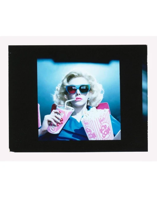 Adult female in blue shirt and 3D glasses drinking from a pink cup and holding popcorn indoors at a movie theater.
