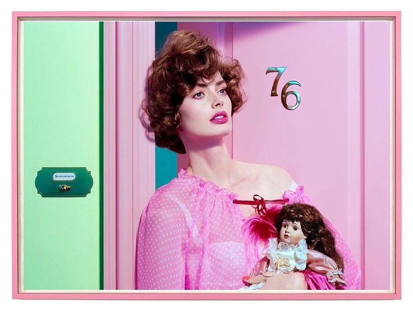 Indoor shot of a woman with brown curly hair in a pink polka dot dress holding a doll, posing in front of a pink door.