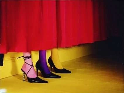 Three women wear high-heeled pumps with colorful tights beneath a red curtain, set against a yellow floor and lit by studio lights.