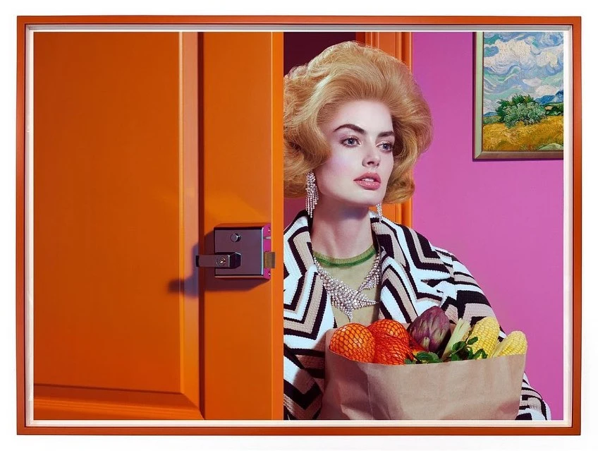 Blonde woman with a 1960s hairstyle holds a bag of produce by an orange door, against a pink wall with a framed painting.
