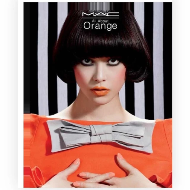 Portrait of a woman with a dark bob haircut wearing an orange top with a large grey bow, posing against a striped backdrop.