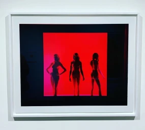 Framed photograph showing three female silhouettes standing against a vivid red backdrop in a gallery setting.