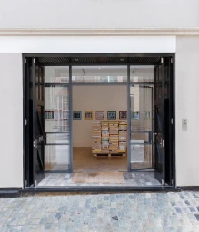 Storefront entrance view showing open dark framed glass doors and a stack of yellow packages inside an art gallery.