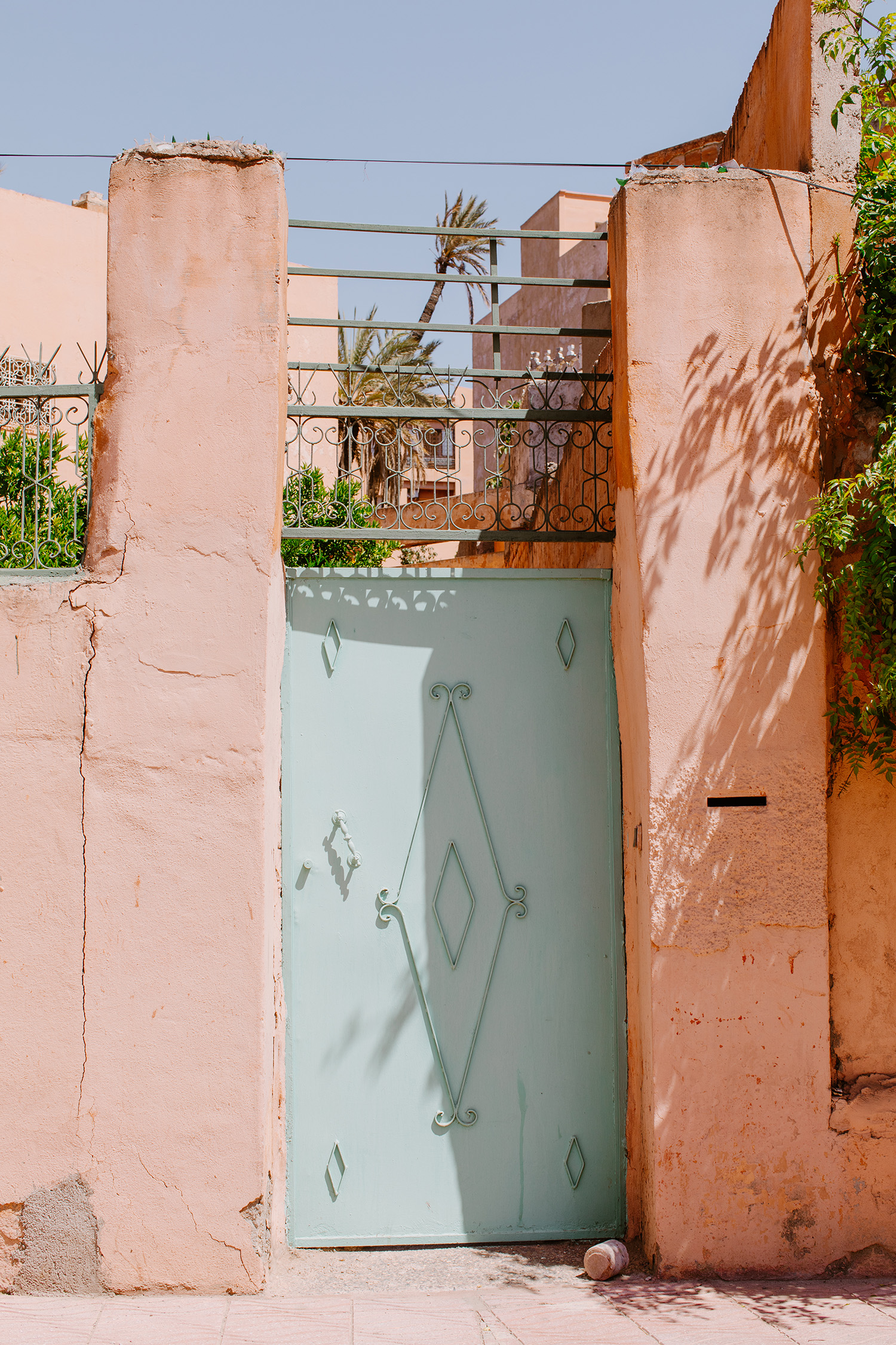 A mint-green gate, adorned with diamond patterns, stands between peach-colored walls in a sun-drenched scene with a palm tree.