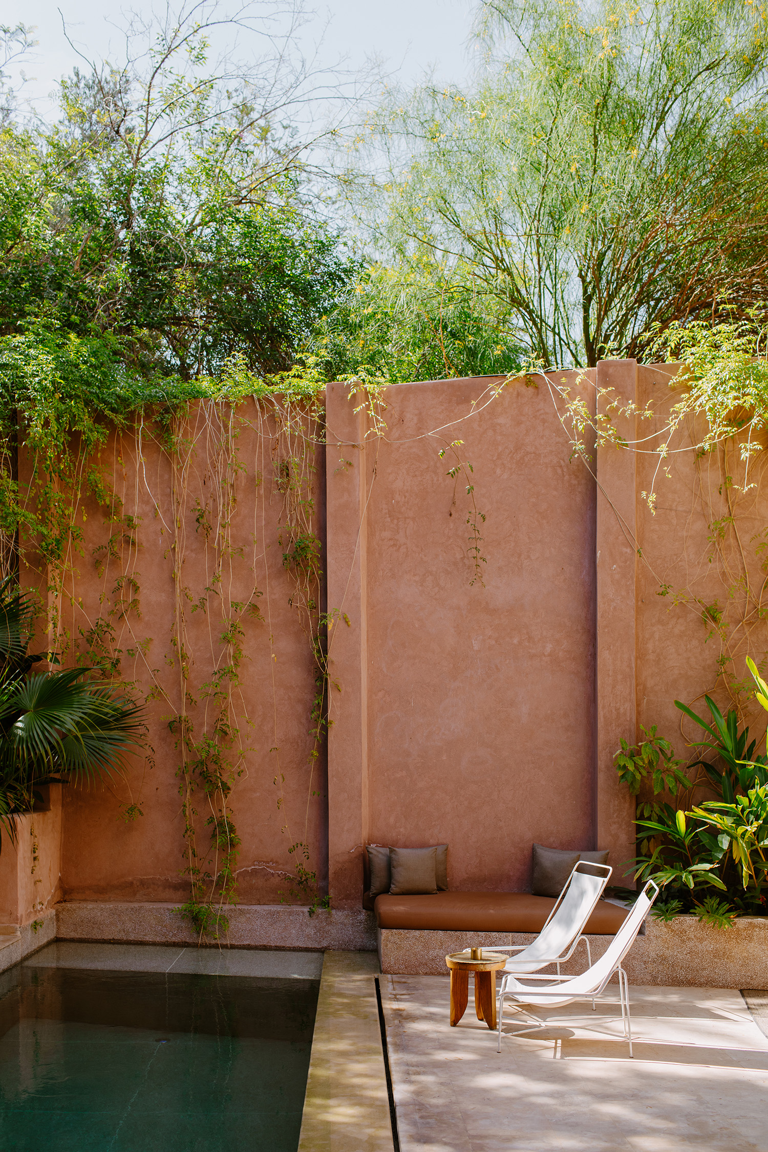 Exterior of a building with a light brown wall, lush green plants, and two white lounge chairs near a pool of water, outdoors.