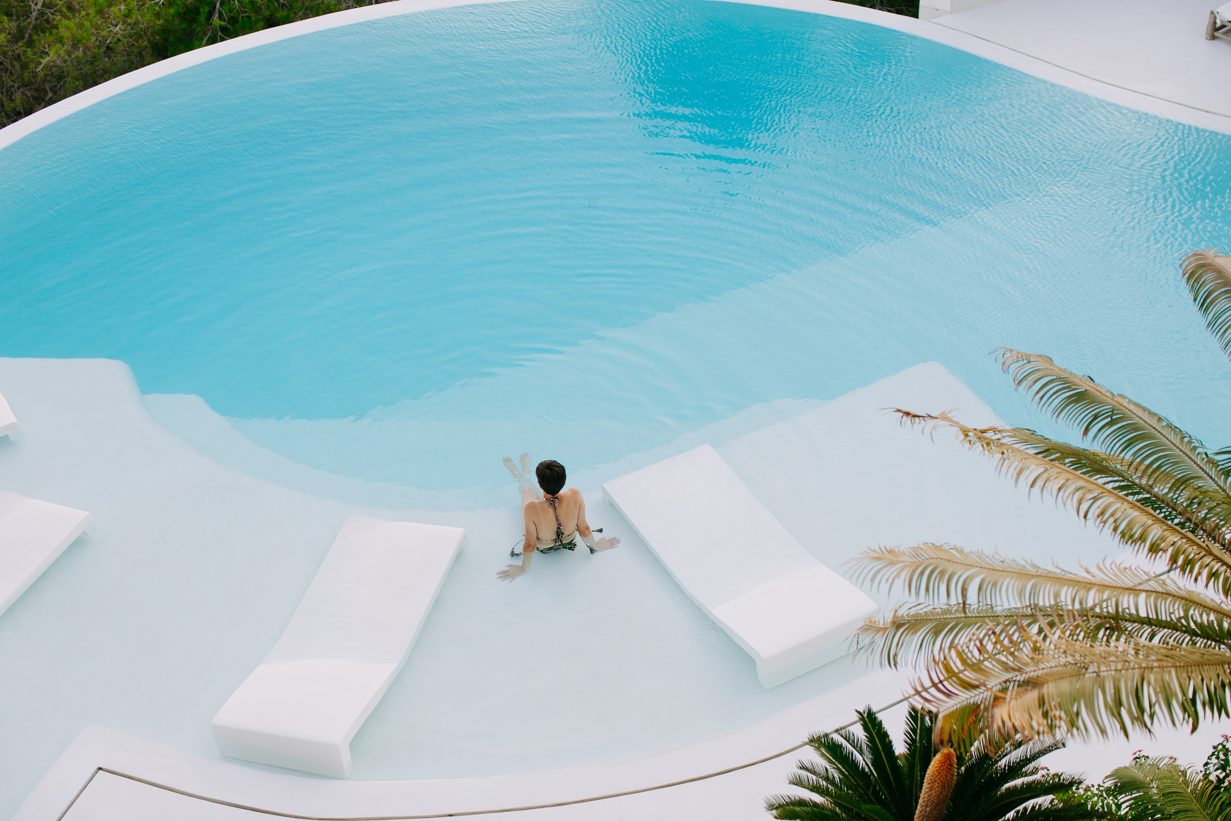 Overhead shot of a woman in a swimming pool with white lounge chairs, surrounded by palm trees, creating a serene vacation scene.