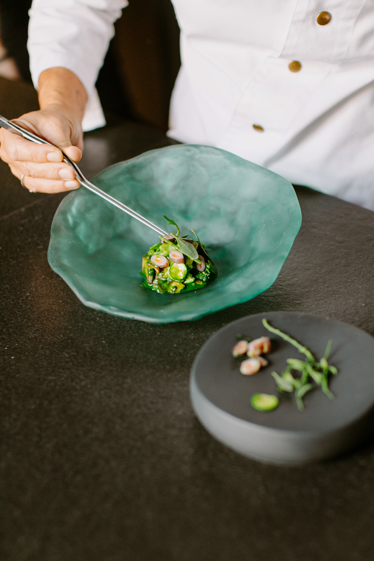 Chef in white uniform delicately places fresh ingredients onto a vibrant green plate on a sleek black surface, professional culinary presentation.