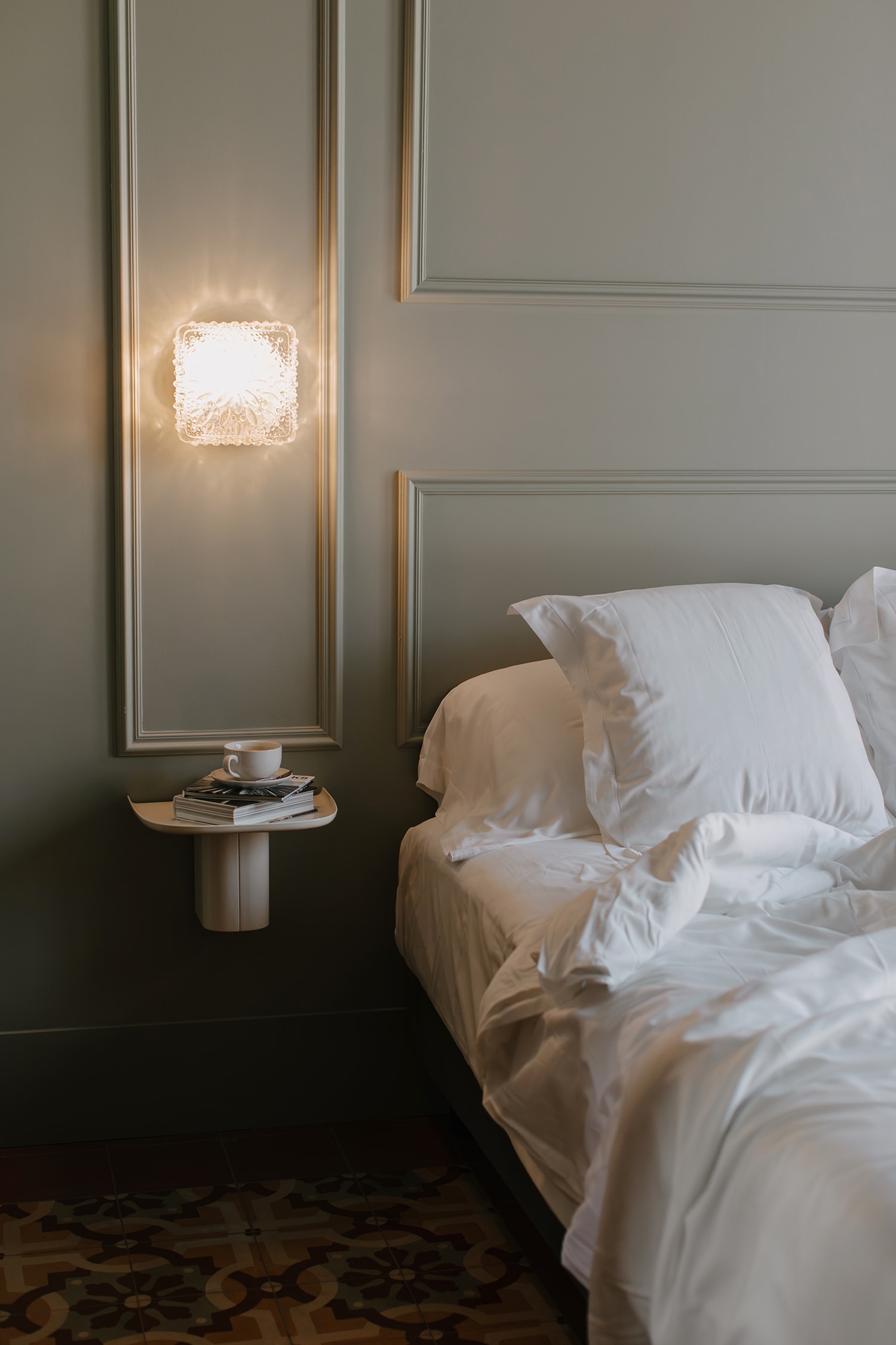 Bedroom interior with a made bed, bedside table with books and a cup, and a decorative light fixture on a gray wall with trim.