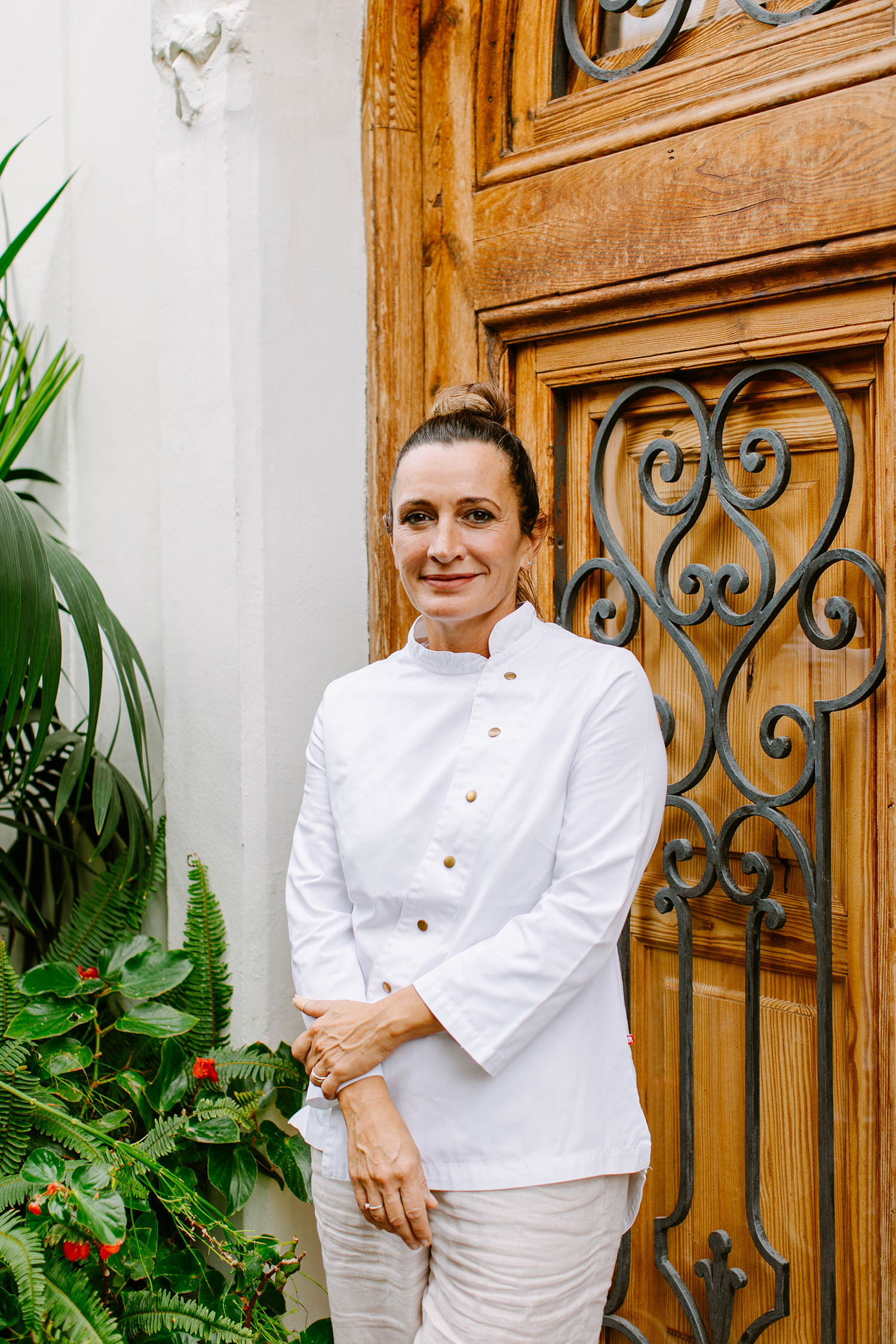 Adult female poses near a wooden door flanked by greenery in a medium shot with natural light, wearing a white top and light pants.
