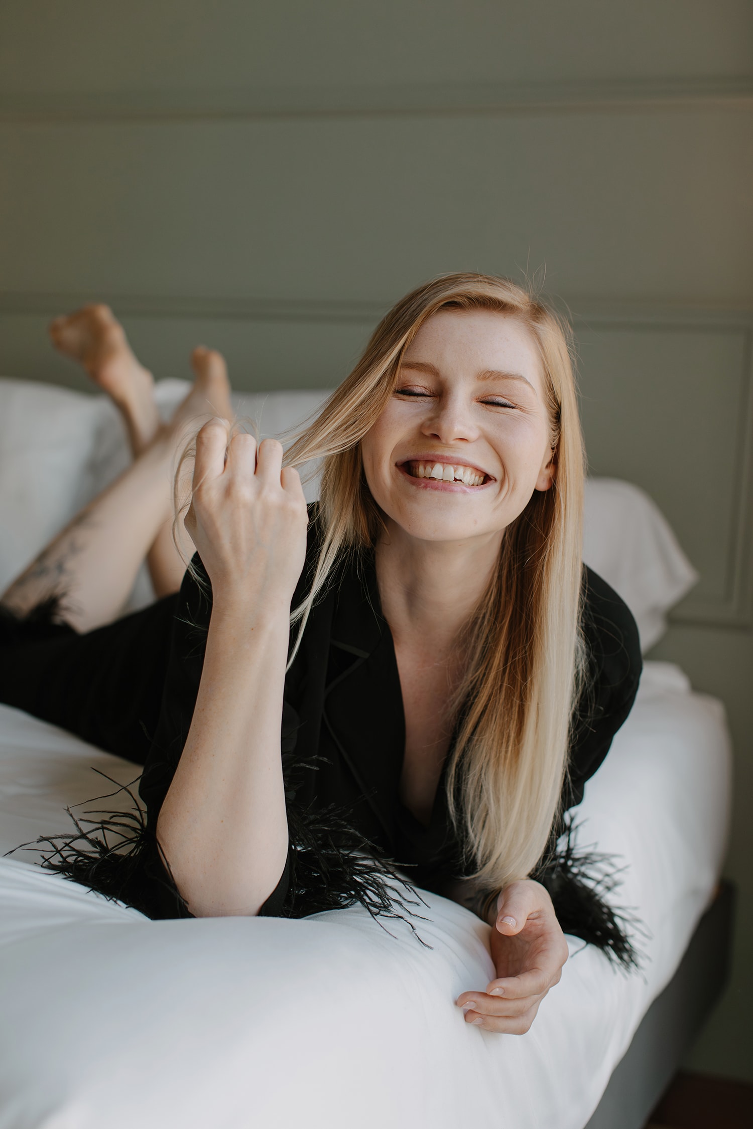 Smiling blonde woman in a black blazer with feathers, lying on a white bed in a room with pale green walls, looking cheerful and relaxed.