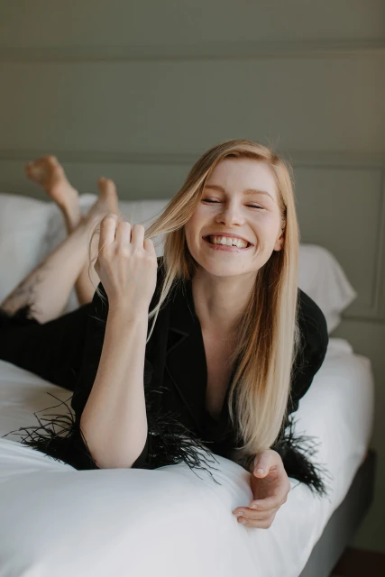 Smiling blonde woman in a black blazer with feathers, lying on a white bed in a room with pale green walls, looking cheerful and relaxed.