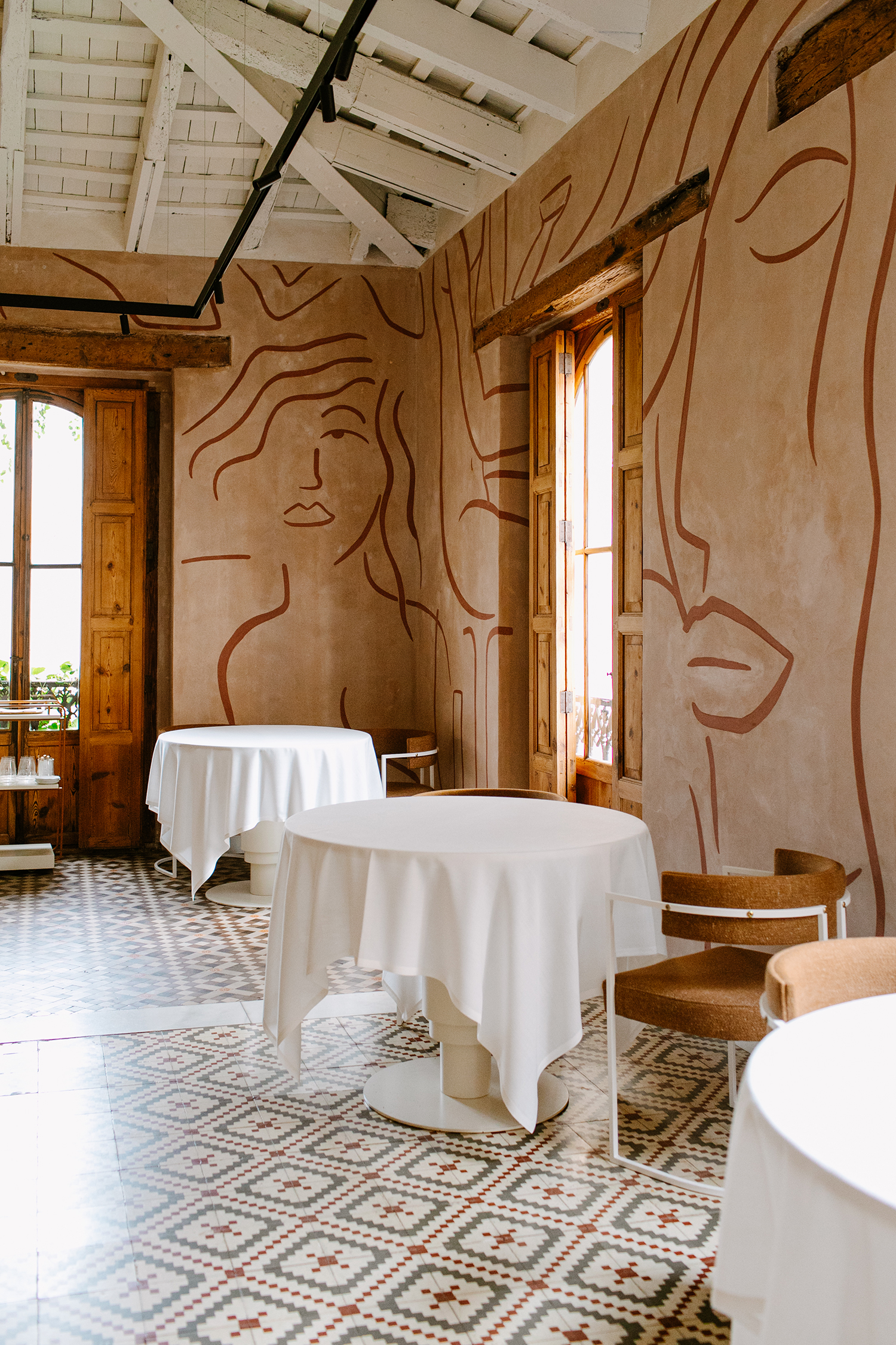 Interior of a restaurant with white tablecloth-covered tables and light brown walls with abstract line art of faces and figures.