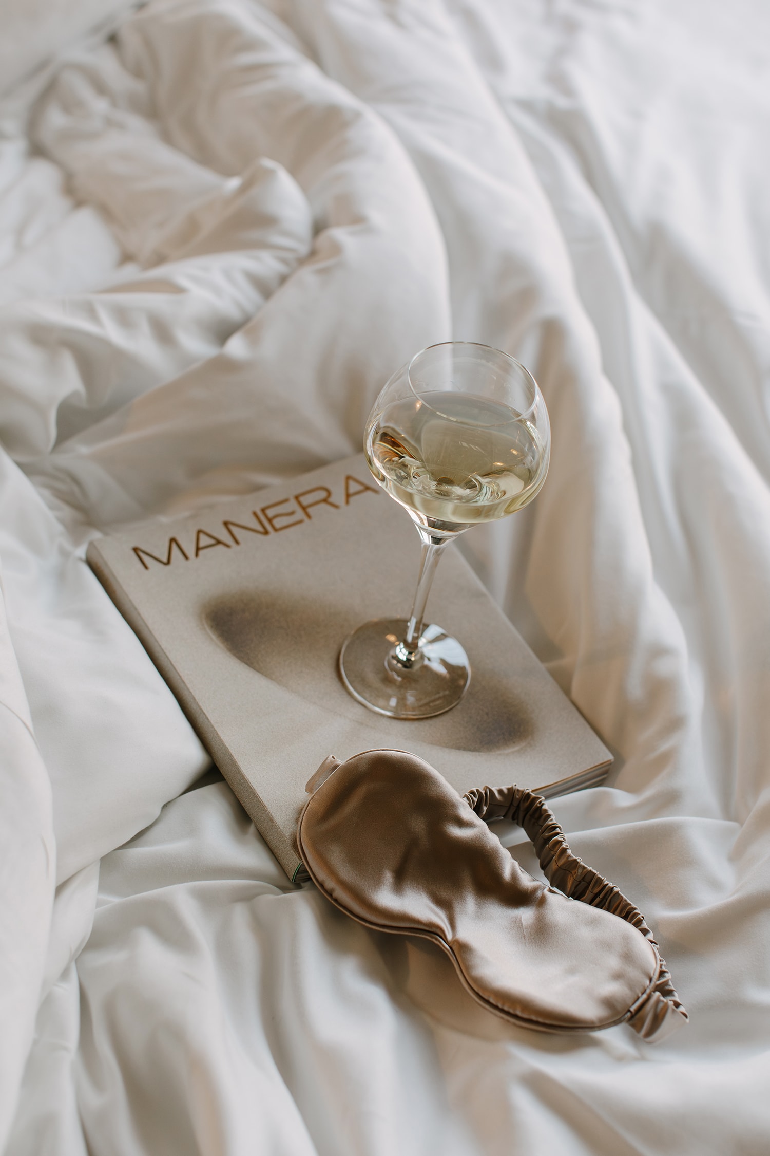 Wine glass on a book on a white bed next to a sleep mask. Relaxing bedroom scene with soft textures and natural light.