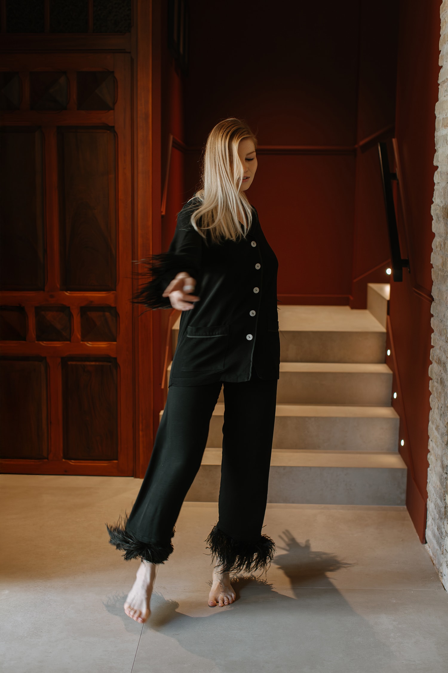 A woman with long blonde hair is shown dancing in a black, feathery pajama set on a gray floor near dark wood and stairs in a brightly lit room.