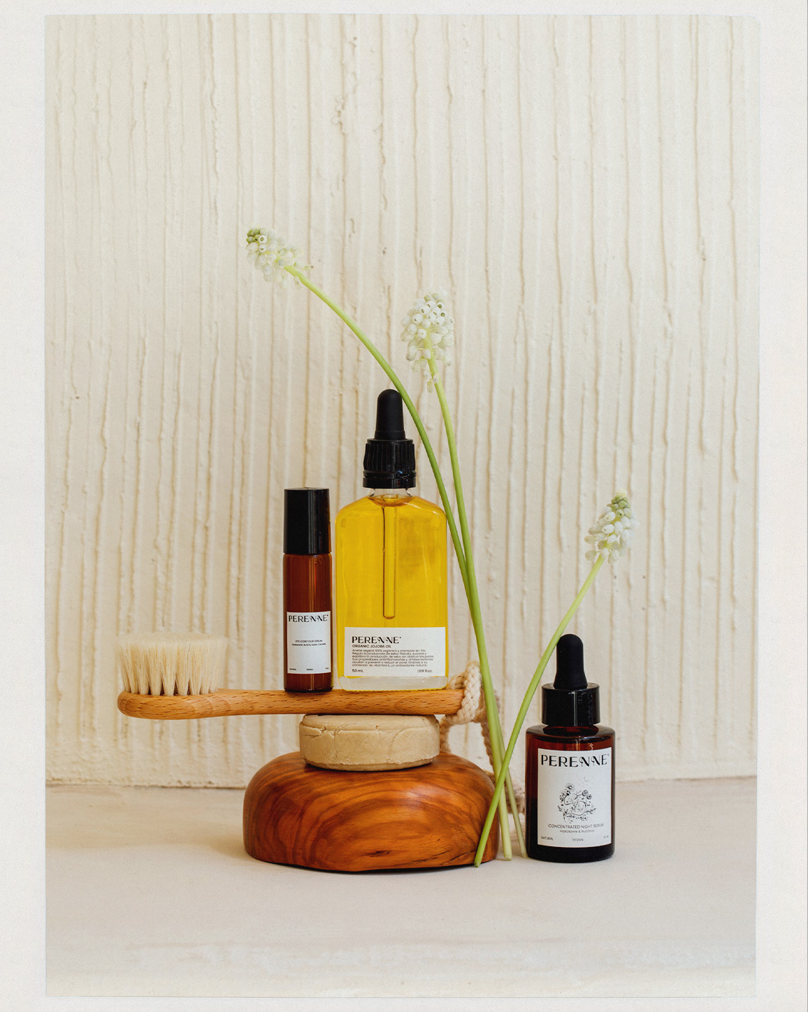 Elevated product shot of PERENAE skincare bottles with floral accents on a wooden platform against a textured white backdrop.