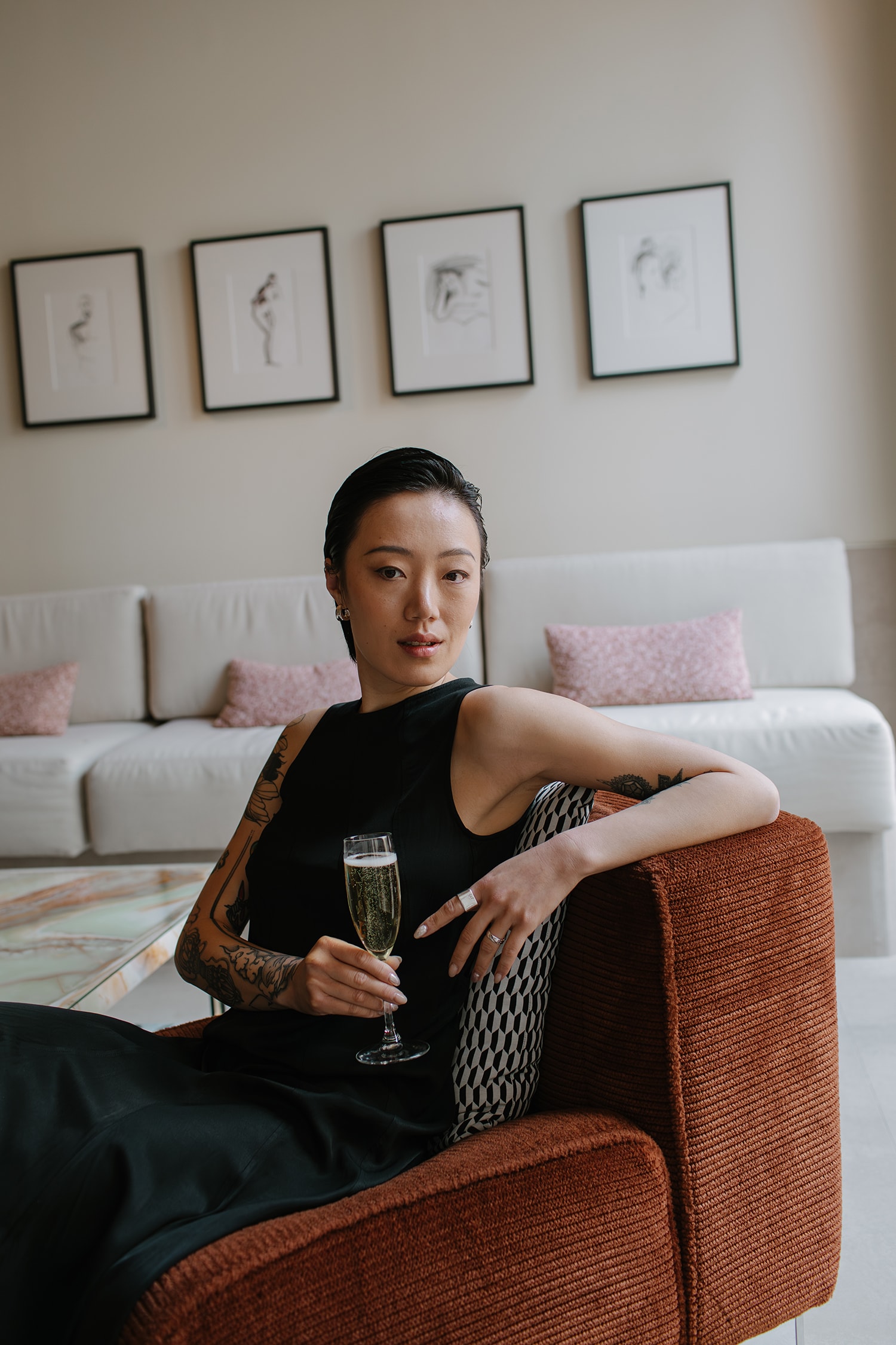 Elegant woman with tattoos in a black dress sitting in an armchair, holding a champagne flute in an indoor living room setting.