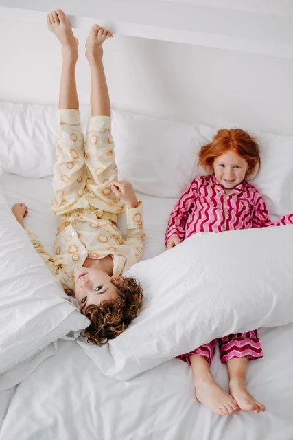 Two children in pajamas are lying on a white bed. One child is upside down with their feet against the wall, while the other smiles.