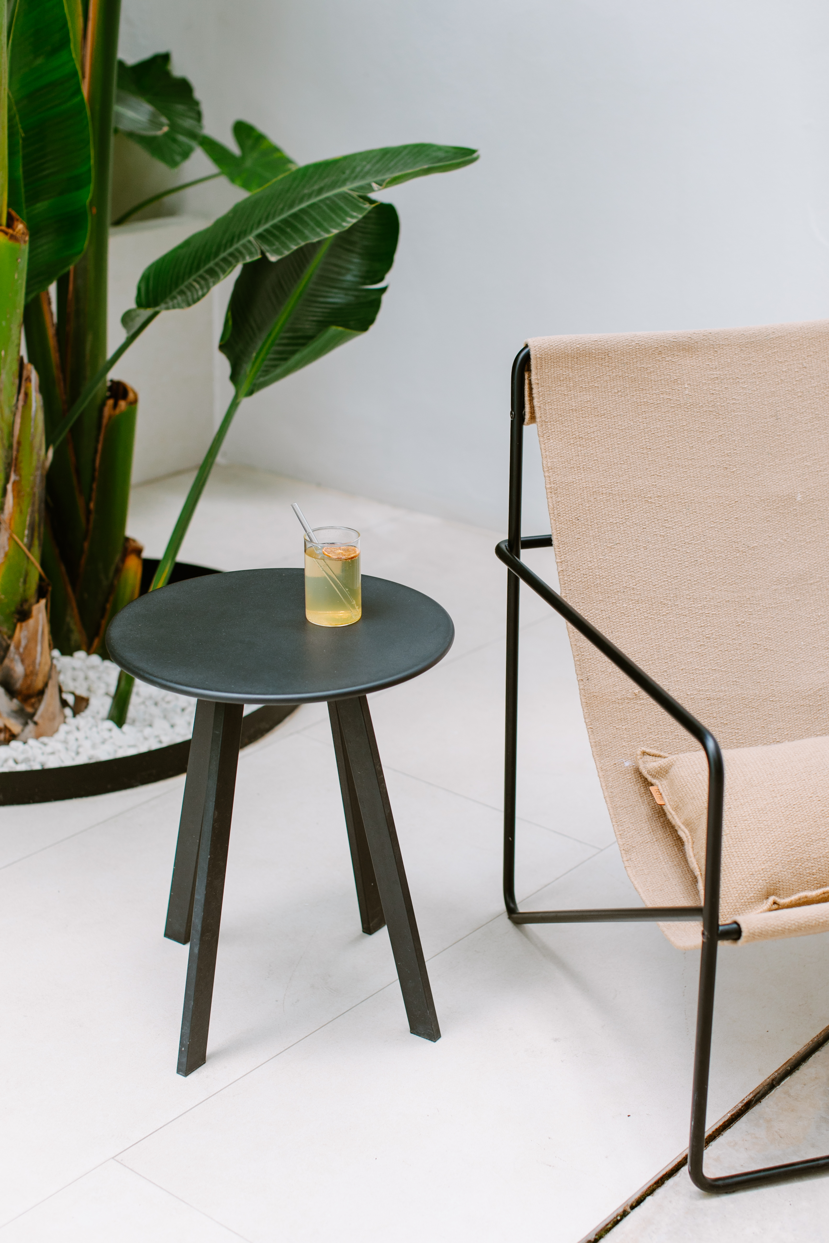 Interior scene with a minimalist chair beside a round side table holding a beverage, near a potted plant against a white wall.