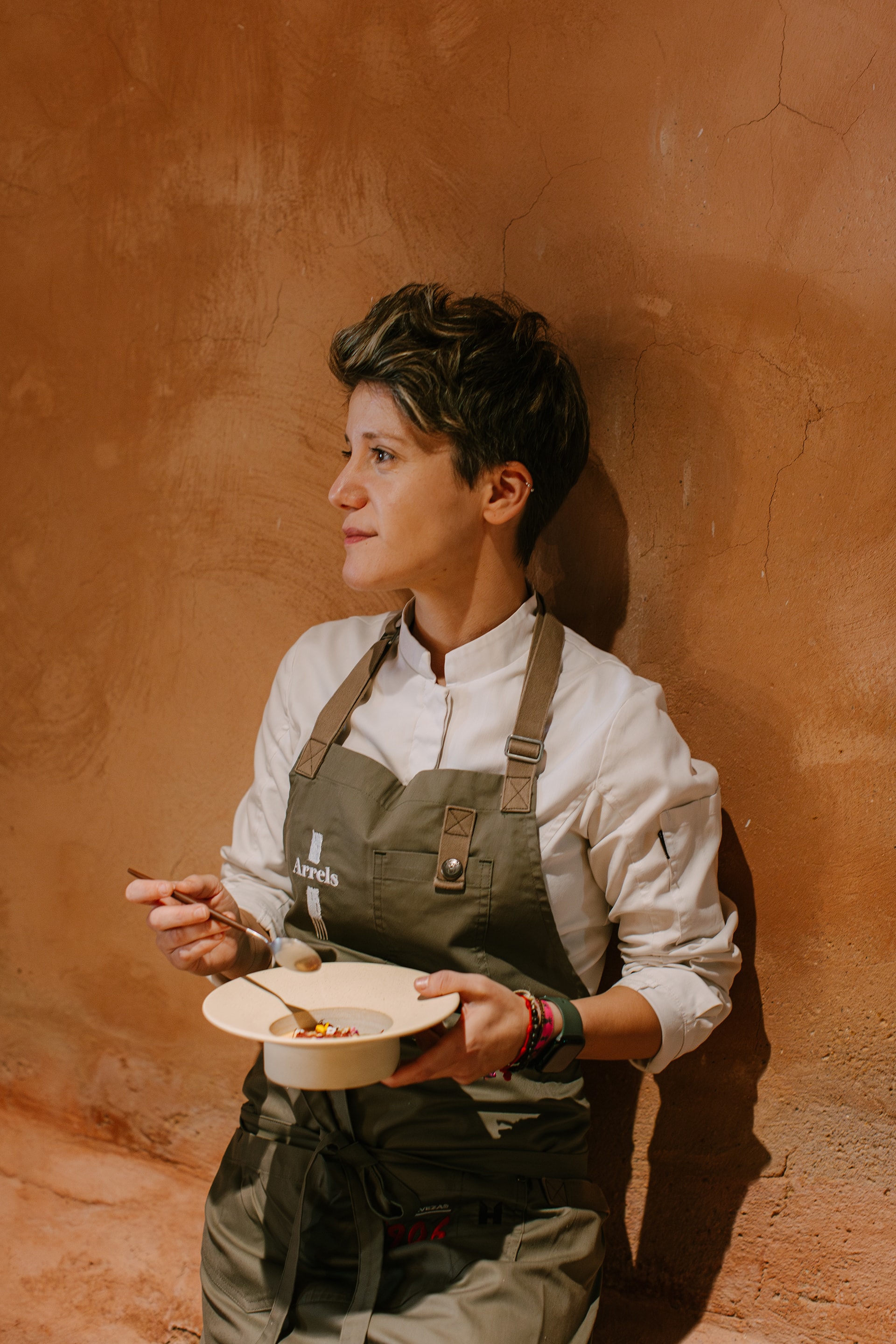Adult female chef in uniform leaning against a textured wall, holding a bowl with spoon, in a warm-toned indoor setting with natural light.