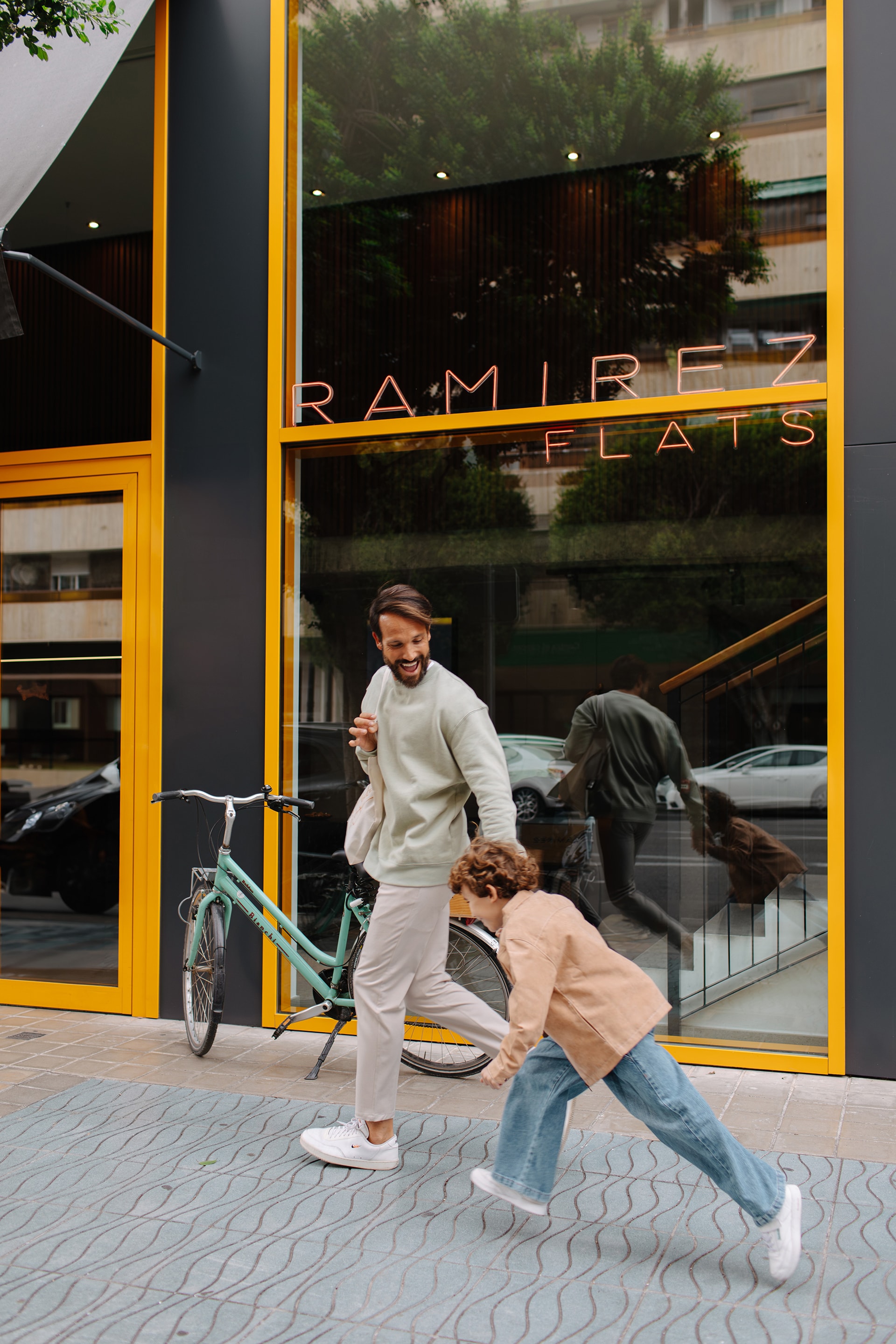 Father and child walking alongside a bicycle in front of a building with large glass windows and yellow accents on a city street.