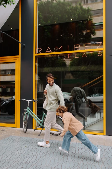 Father and child walking alongside a bicycle in front of a building with large glass windows and yellow accents on a city street.