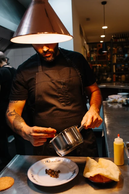 Professional chef prepares food in a restaurant kitchen. Man plating ingredients with metal pot and plate under bright copper light.