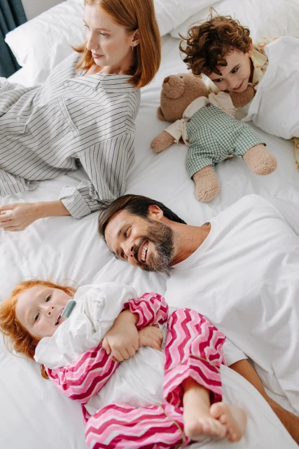 A family of four, including mother, father, son, and daughter, lie together on a white bed in their pajamas, smiling and relaxed.