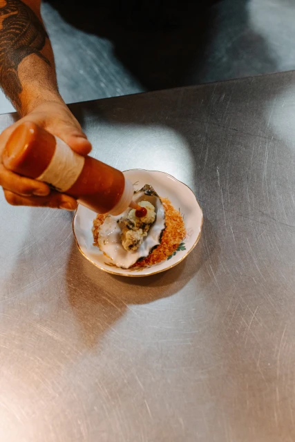 Overhead shot of a chef adding sauce to a raw oyster on a decorative plate, resting on a stainless steel surface in a restaurant.
