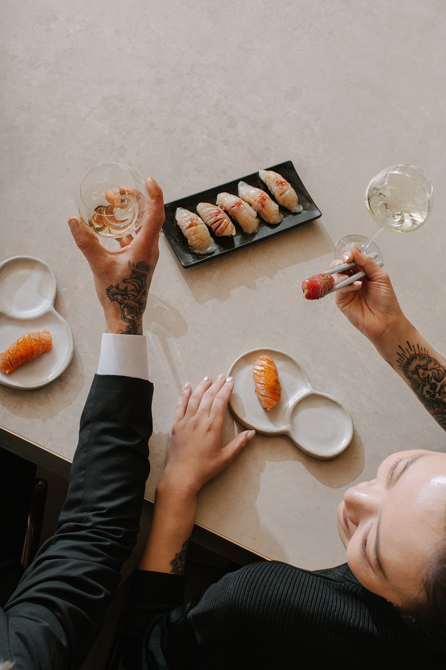 Overhead shot of two people at a table with sushi, one person holding chopsticks with sushi, the other holding a glass of wine.