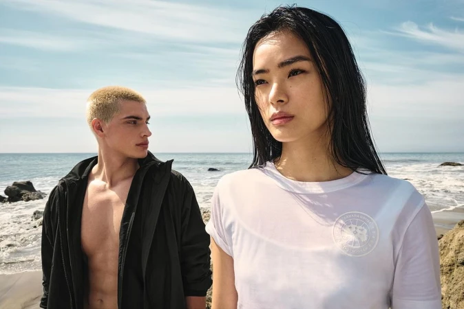 Long-haired Asian woman in white t-shirt and short-haired blonde man in black jacket standing on a beach by the ocean