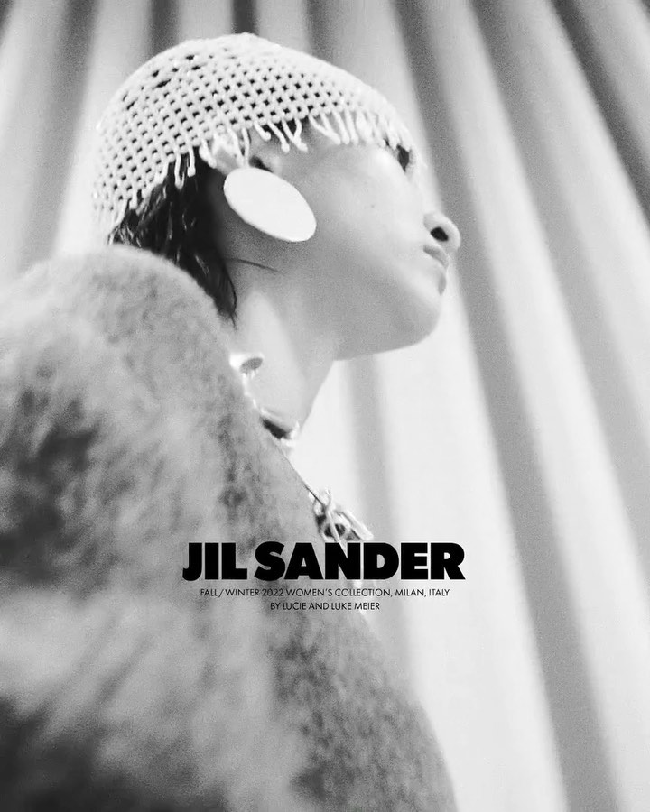 Black and white fashion portrait of a person wearing a crocheted hat, large disc earrings, and a wool coat in front of draped curtains.