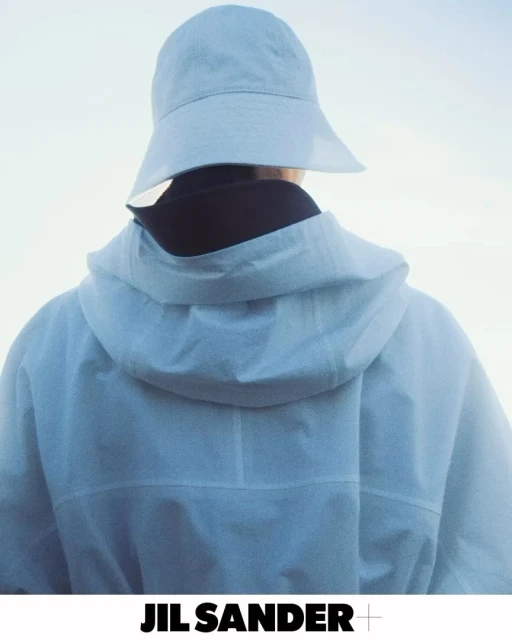 Person from behind wearing a light blue jacket and hat against a light sky, with JIL SANDER logo at the bottom of the frame.