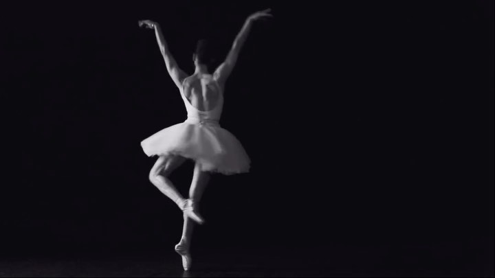 Back view of a female ballet dancer in white tutu en pointe with arms raised against a dark background in a black and white photograph.