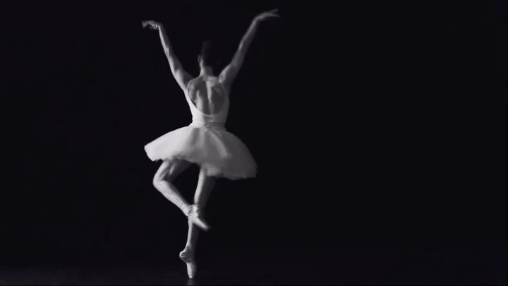 Back view of a female ballet dancer in white tutu en pointe with arms raised against a dark background in a black and white photograph.