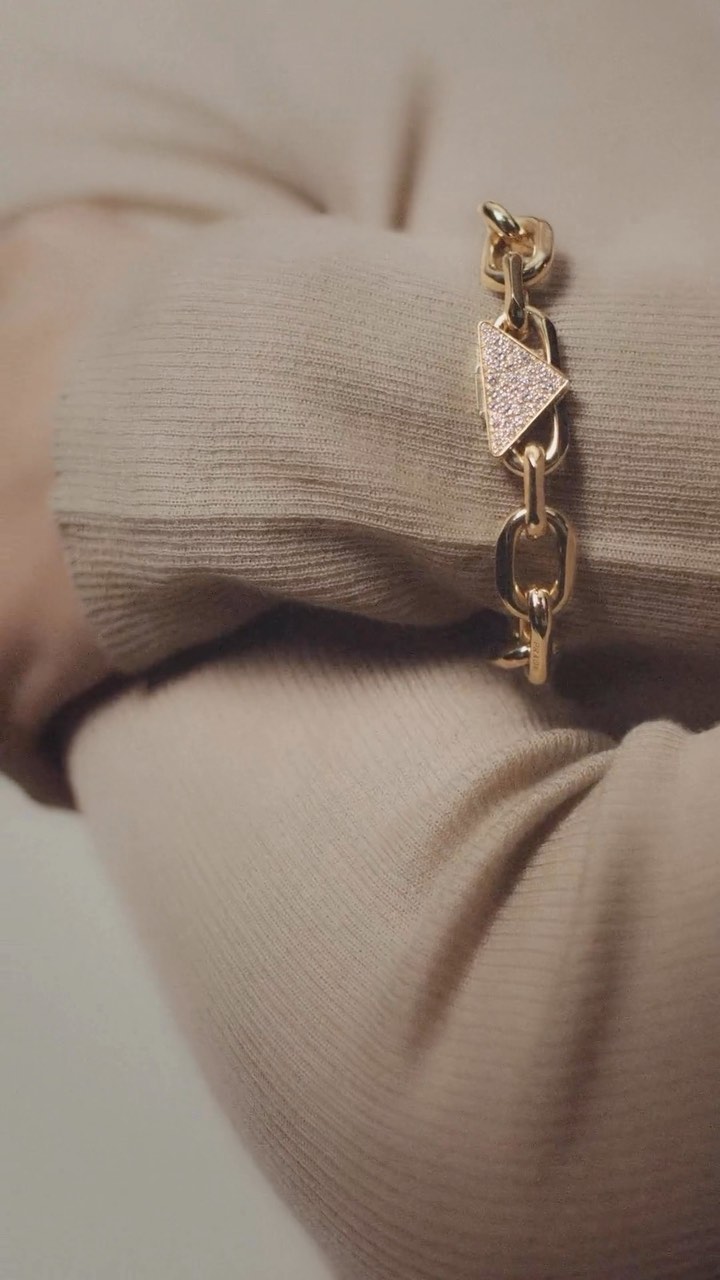 Close-up of a gold chain-link bracelet with a triangle charm accent adorning a person's wrist, set against a neutral sweater background.