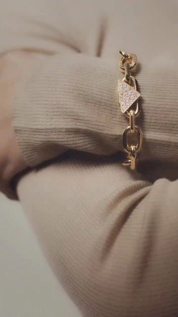 Close-up of a gold chain-link bracelet with a triangle charm accent adorning a person's wrist, set against a neutral sweater background.
