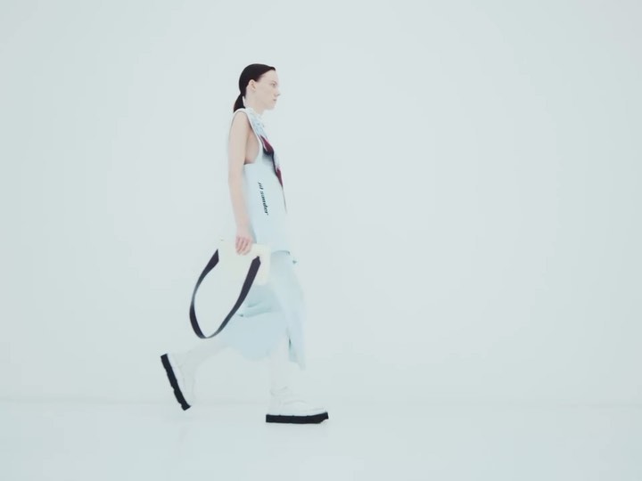 Woman in modern light blue fashion, walking in a minimal white studio, carrying a white tote bag with black straps.