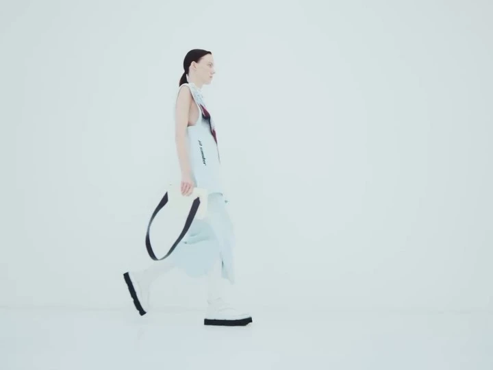 Woman in modern light blue fashion, walking in a minimal white studio, carrying a white tote bag with black straps.