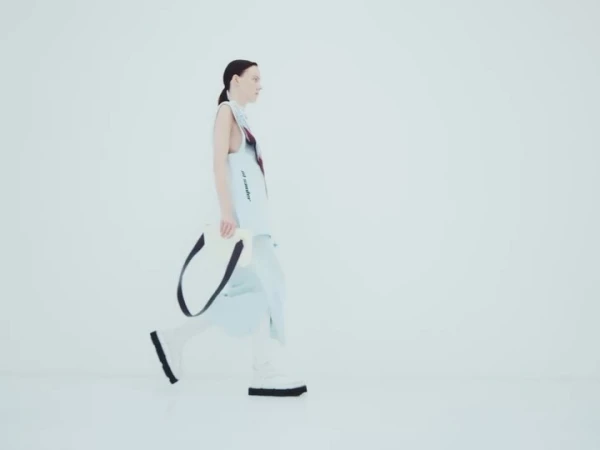 Woman in modern light blue fashion, walking in a minimal white studio, carrying a white tote bag with black straps.