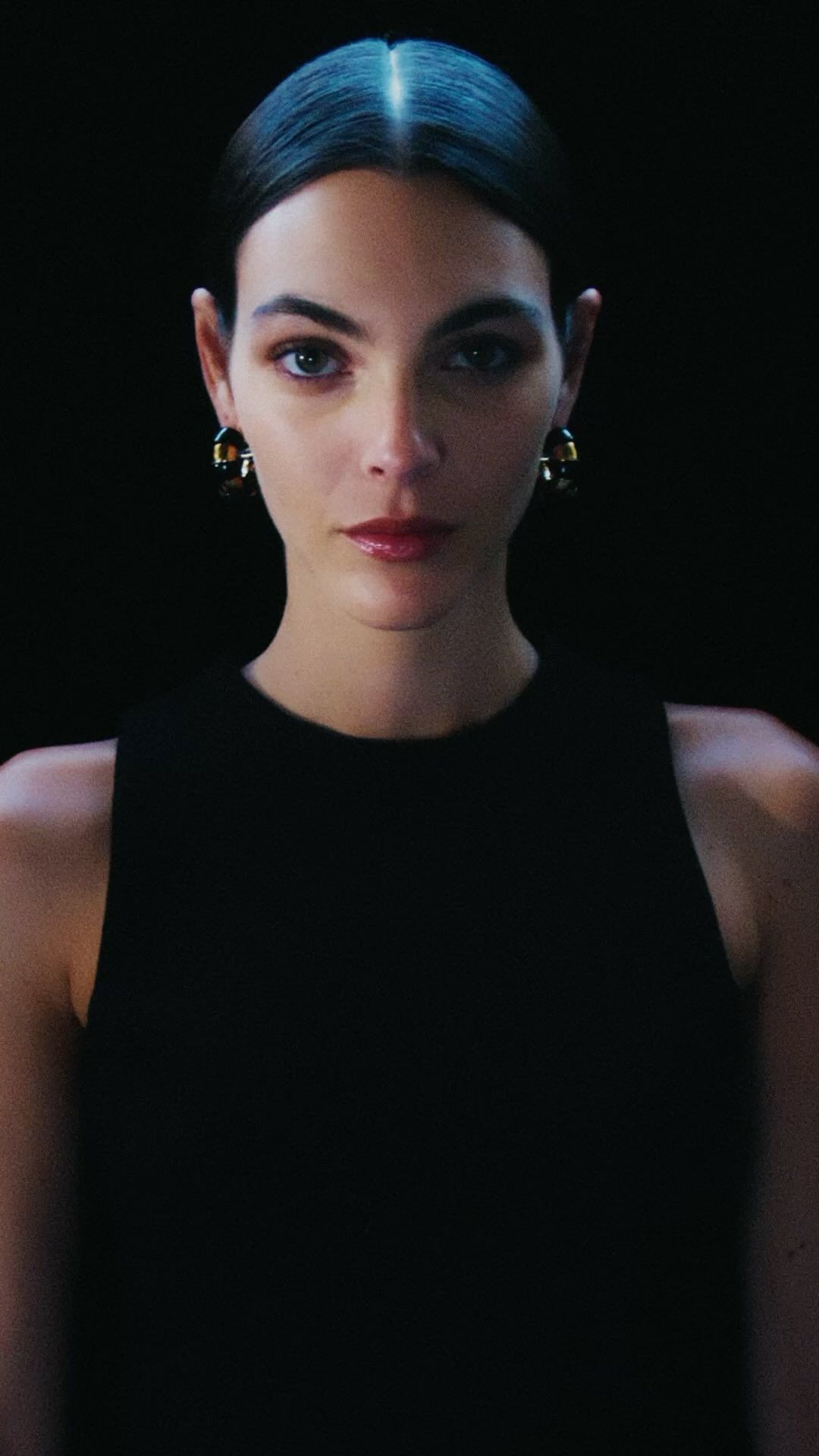 Glamorous portrait of a woman with dark hair wearing a black top and gold earrings against a black background in a photo studio.
