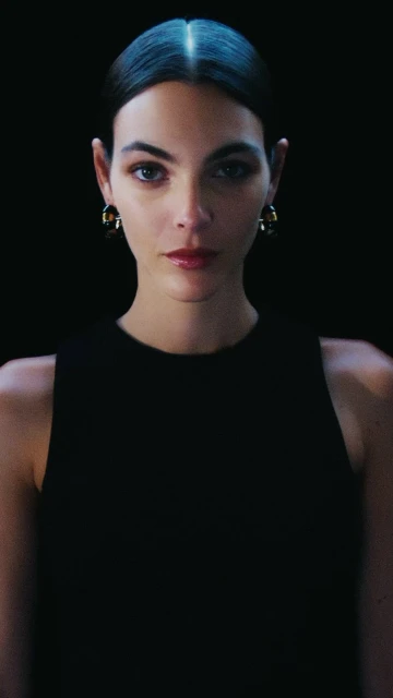Glamorous portrait of a woman with dark hair wearing a black top and gold earrings against a black background in a photo studio.