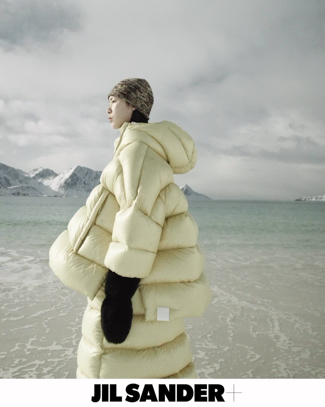 Fashion shot of Asian woman wearing a large, hooded puffer coat and knit beanie, standing on a beach with mountains in the background.