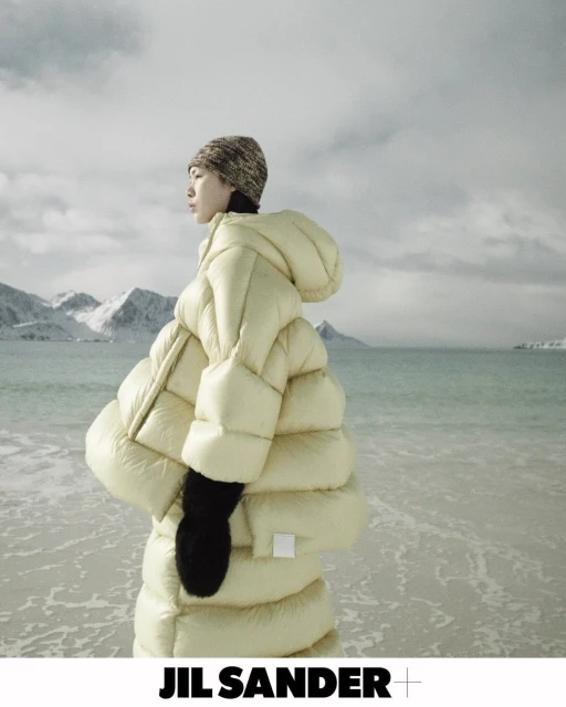 Fashion shot of Asian woman wearing a large, hooded puffer coat and knit beanie, standing on a beach with mountains in the background.