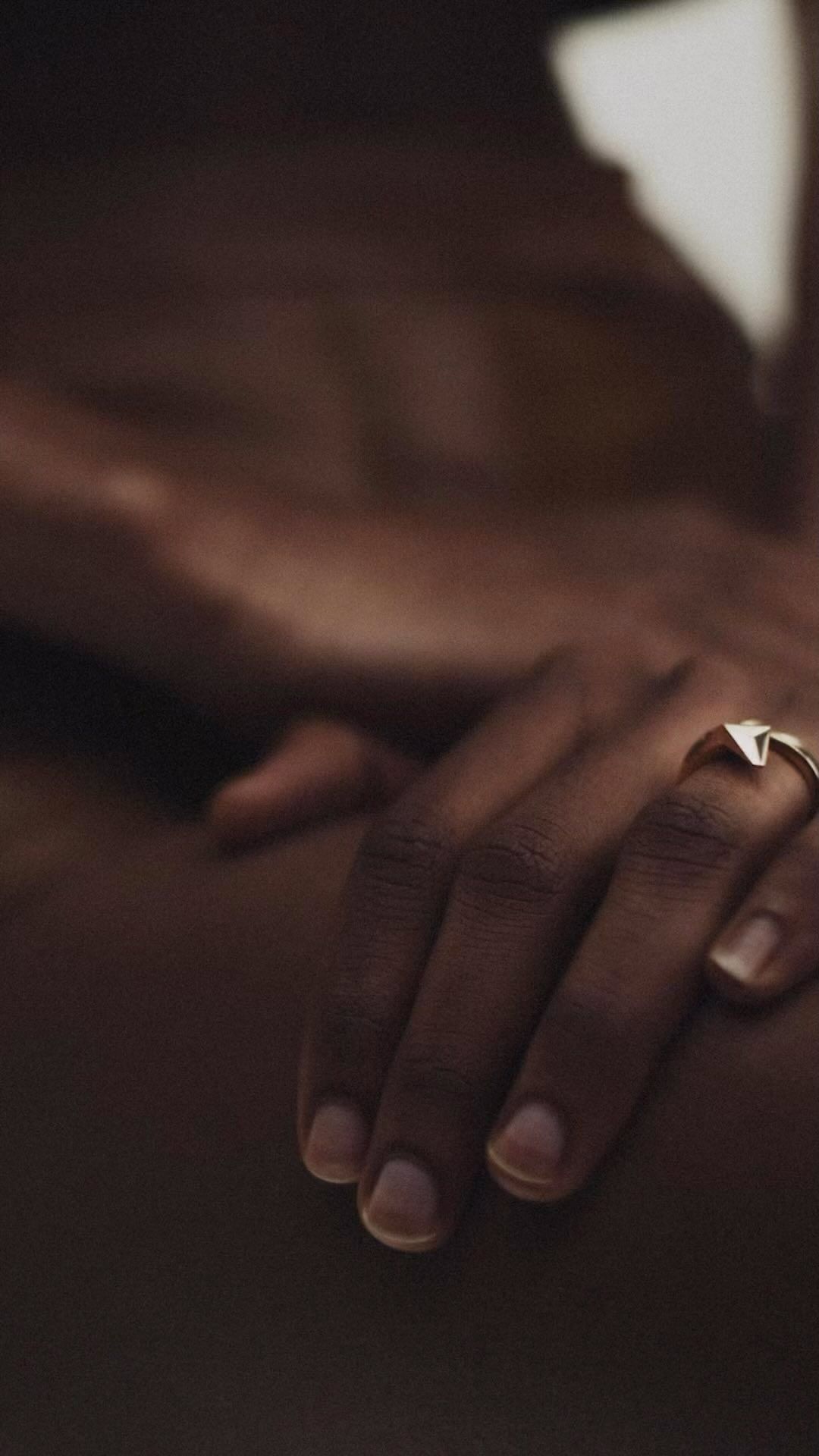 Close-up studio shot of a dark-skinned person's hands with a gold ring, bathed in warm light, showcasing minimal nude skin.